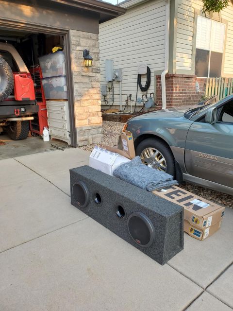 1996 Ford Bronco - A gray Subaru Outback is parked in a driveway next to a subwoofer box and car parts, with a portion of a Jeep visible in the garage.