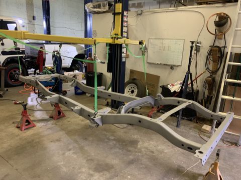 1979 Jeep CJ5 - A vehicle frame is suspended by a yellow lift in a workshop, with a white truck visible in the background.