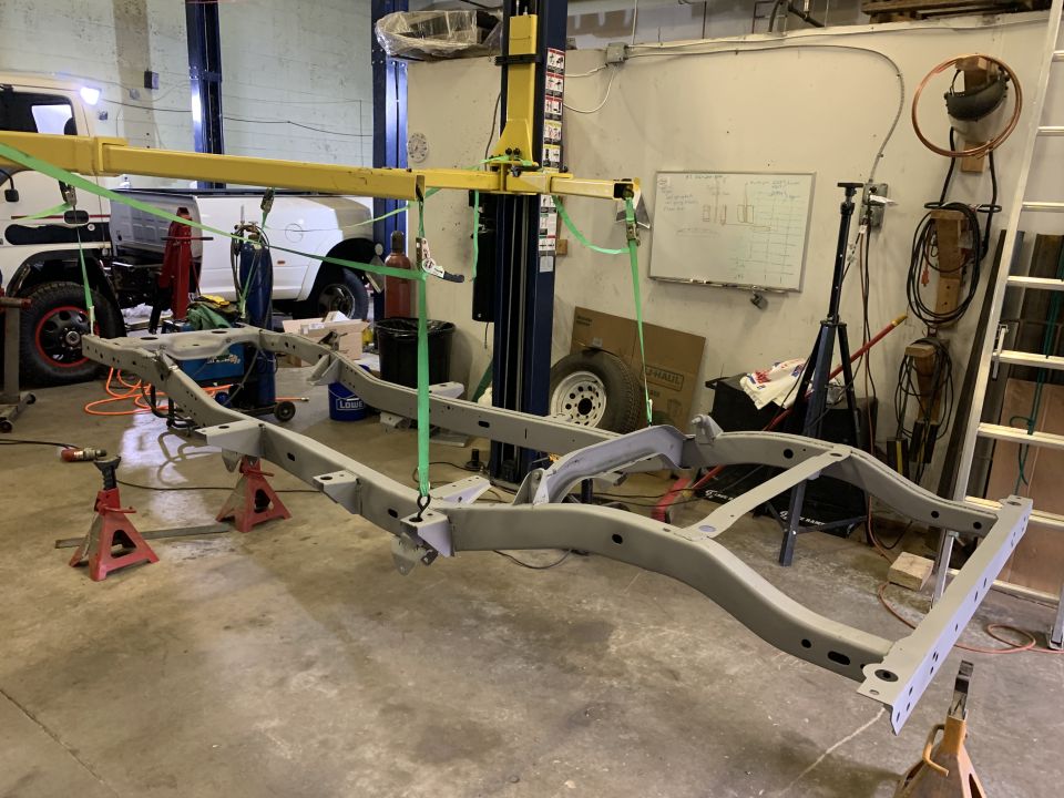 vehicle frame suspended by yellow lift in garage, white truck in background