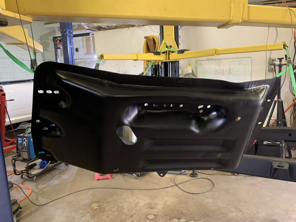 Black metal panel, possibly a fender or body panel, suspended by green straps from a yellow overhead lift in a workshop