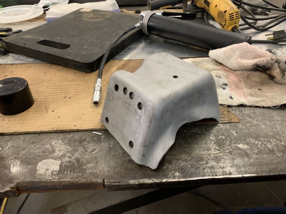 Gray metal bracket with multiple bolt holes and a connected grease gun on a workbench