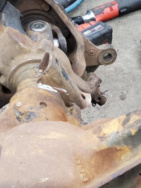 1996 Ford Bronco - Close-up of a heavily rusted suspension part, likely from a vehicle undergoing modification or repair, with a power tool visible in the bac…