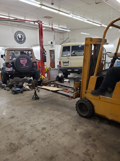 1996 Ford Bronco - A garage scene shows a Jeep Wrangler and a Toyota Land Cruiser with mechanical parts being moved by a forklift.