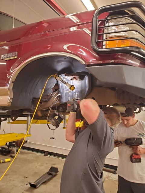 1996 Ford Bronco - Two people are working on the underside of a red Ford Bronco, which is raised on a lift.