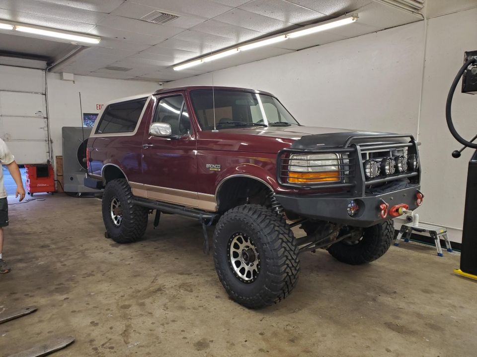Maroon Ford Bronco with a white top, lifted suspension, and off-road tires. Modified front bumper with lights