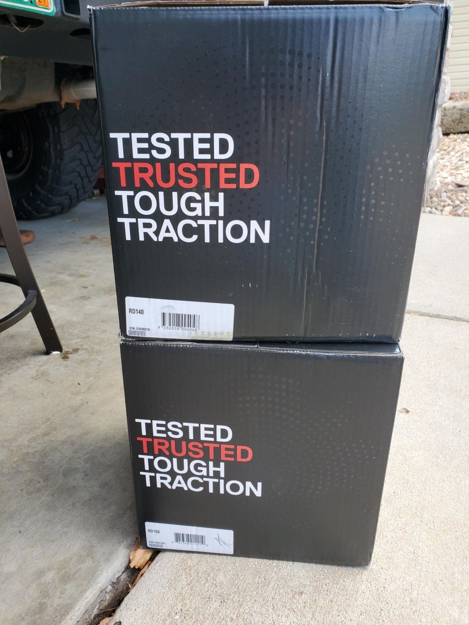 Two black boxes stacked, each saying 'TESTED TRUSTED TOUGH TRACTION'. Part of a vehicle tire and bumper visible to the left