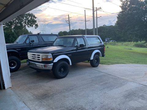 1996 Ford Bronco - Two black Ford Broncos are parked outside on a paved surface near a garage.