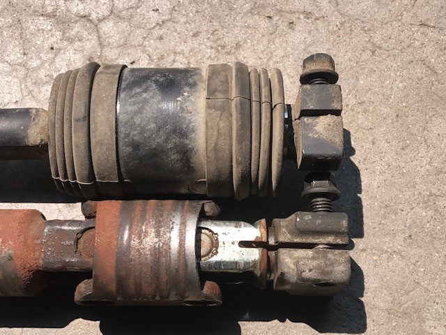 Two shock absorbers, one with a cracked rubber boot and the other showing rust, are placed side-by-side on a concrete surface
