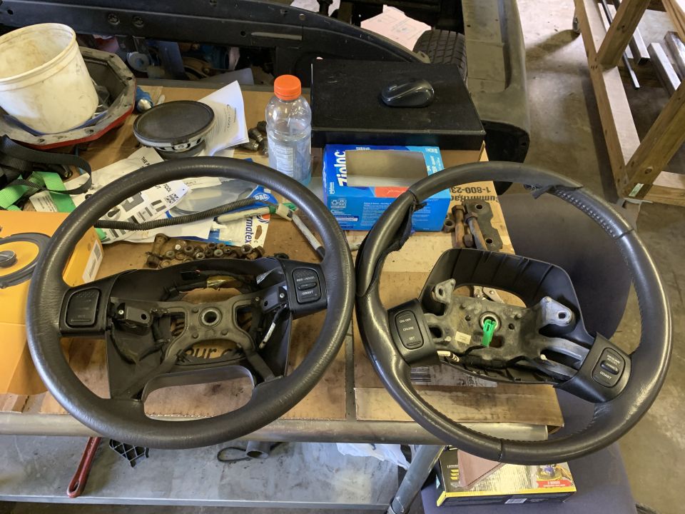 Two car steering wheels are on a workbench with various parts and a bottle of water
