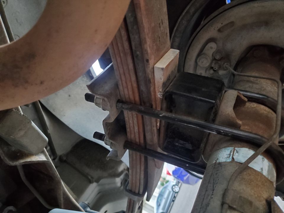 Close-up of a vehicle's leaf spring and suspension components. A wooden block is visible near the spring