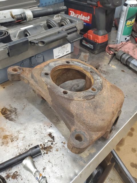 1996 Ford Bronco - A heavily rusted steering knuckle sits on a workbench next to various tools and a Duralast battery.