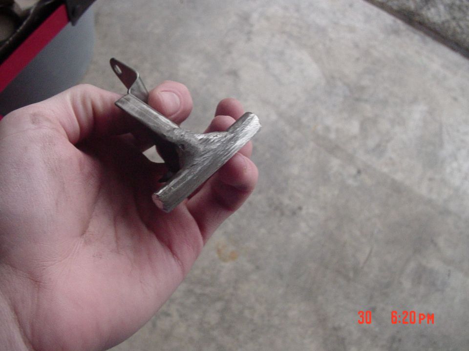 Hand holding a small welded T-shaped metal bracket with one mounting hole over a garage floor