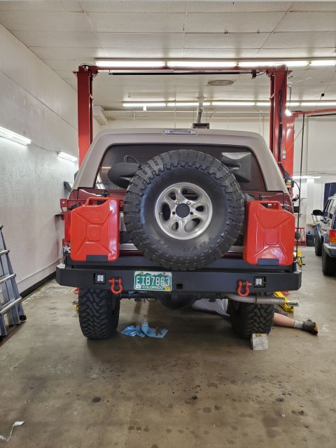 1996 Ford Bronco - A 1996 Ford Bronco is shown from the rear, equipped with a spare tire, dual jerry cans, and a custom bumper, while a mechanic works underne…