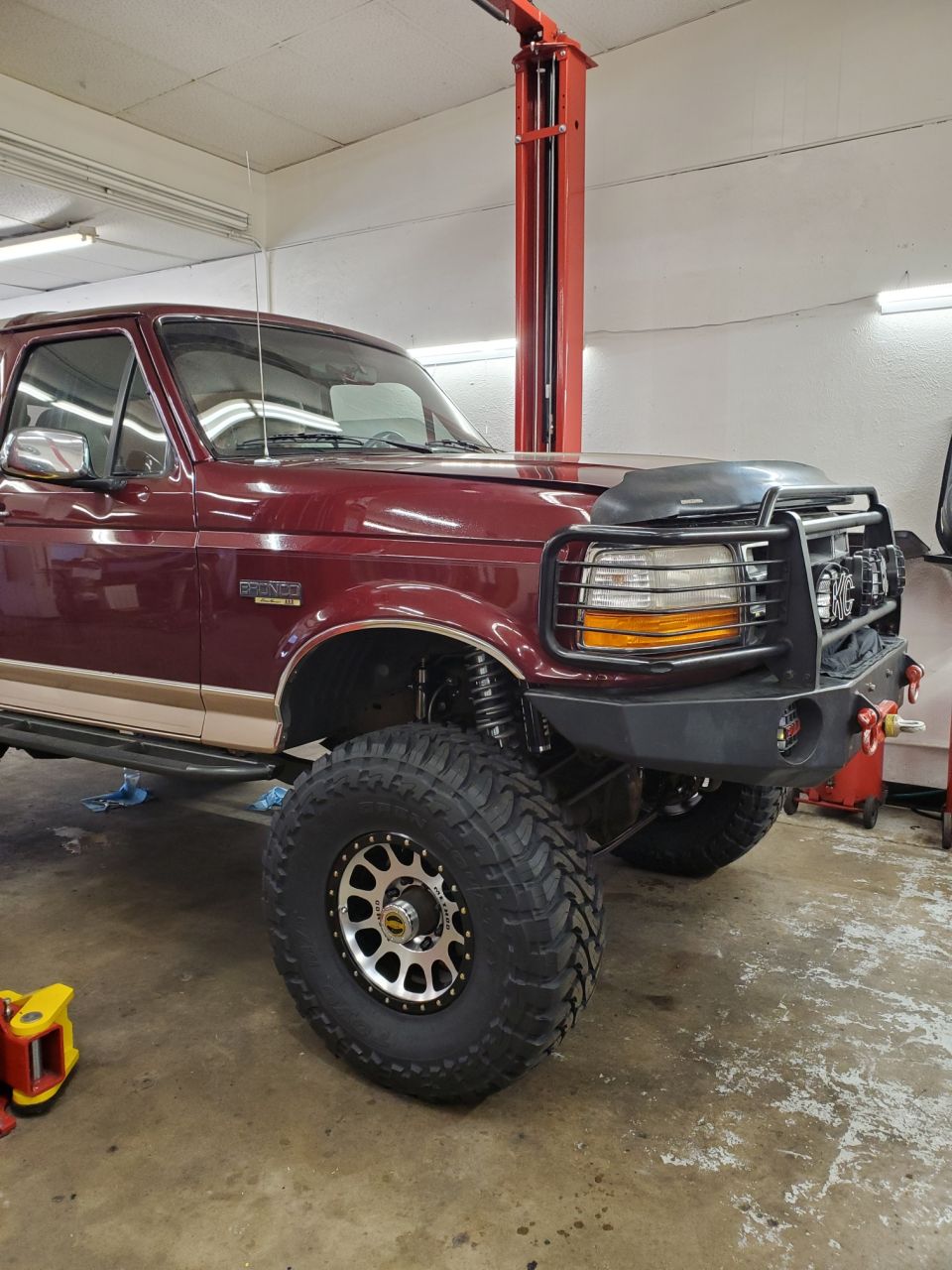 Maroon Ford Bronco lifted with large tires and aftermarket bumper, on a lift in a garage