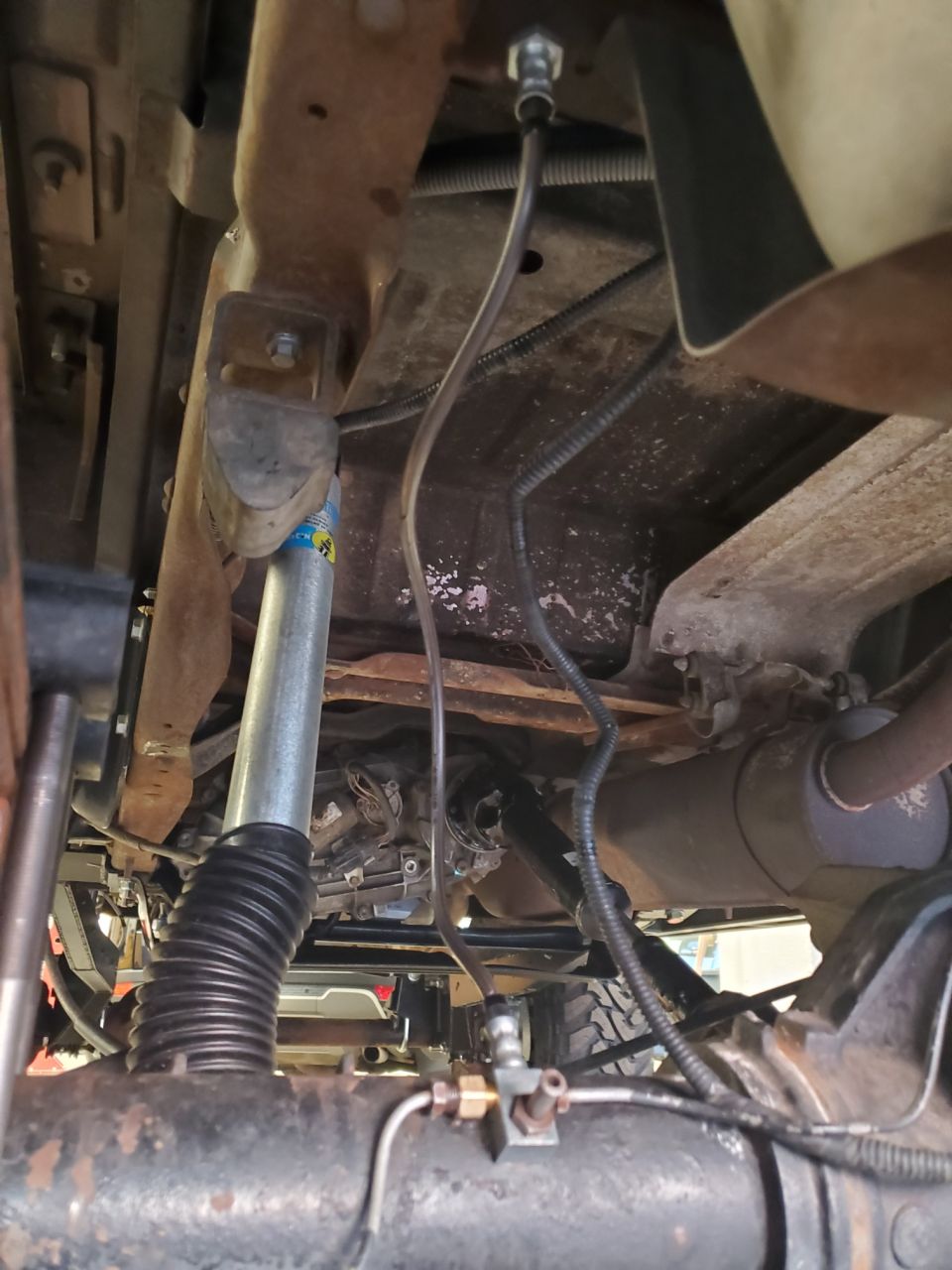 Underside view of a truck with a Bilstein shock absorber and custom suspension components. Shows frame, exhaust, and drivetrain parts