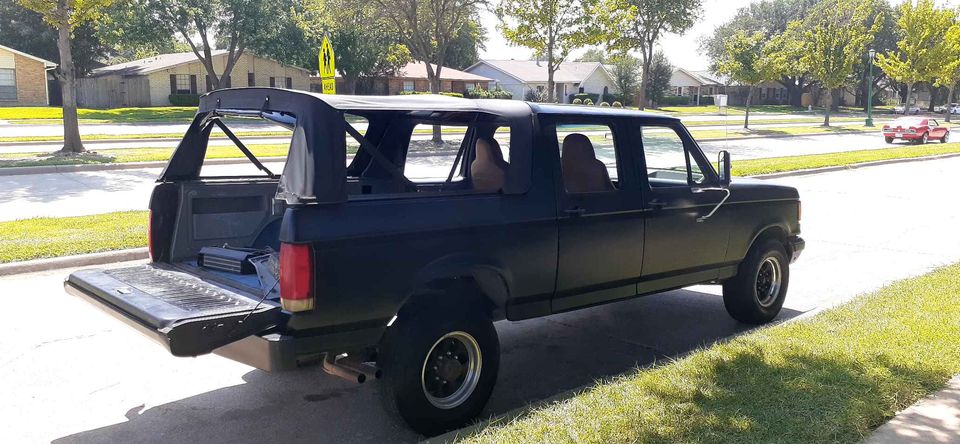 Matte black Ford Bronco with tailgate down, amplifier in cargo area, soft top removed, parked on residential street.