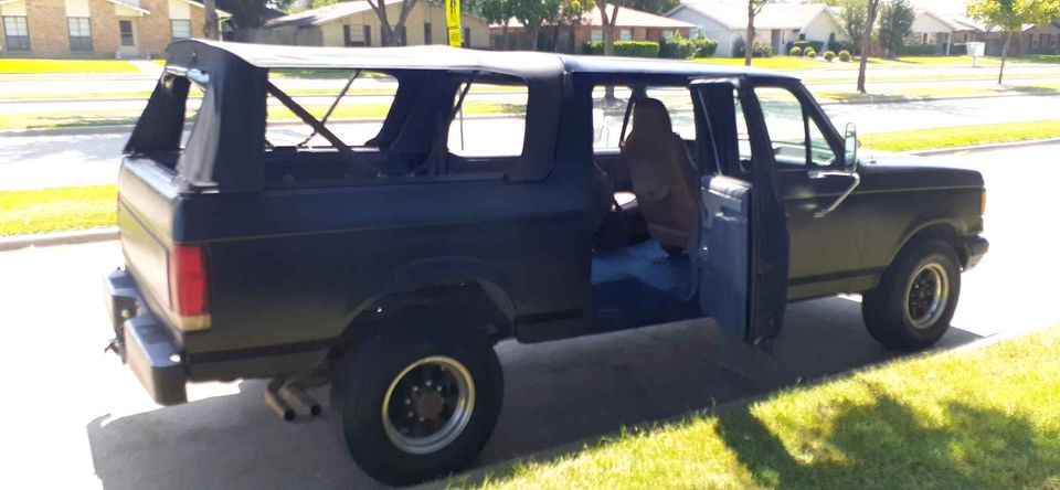 Dark blue Ford Bronco with soft top frame, driver side door open, parked on street.