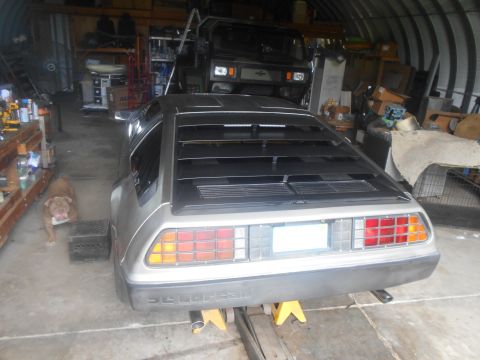 1992 Ford Bronco - The rear view of a silver DeLorean DMC-12 is shown in a cluttered garage. A tan dog stands to the left of the car.