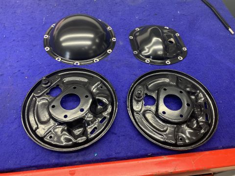 1979 Jeep CJ5 - Two black differential covers and two black brake backing plates are laid out on a blue surface. The differential covers are domed and flat…