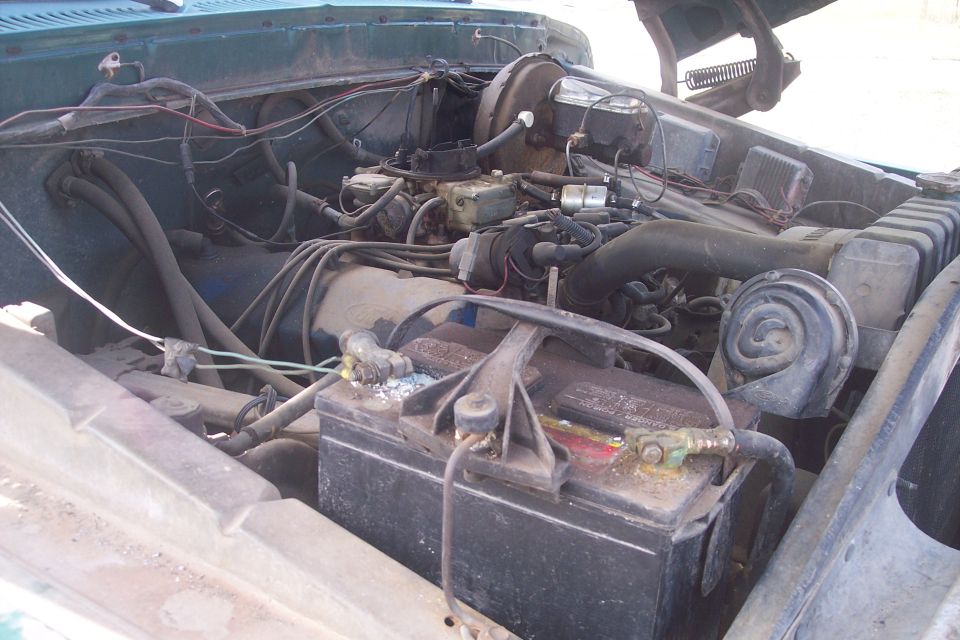 Engine bay of a 1977 Ford F-150 showing the battery, carburetor, and hoses