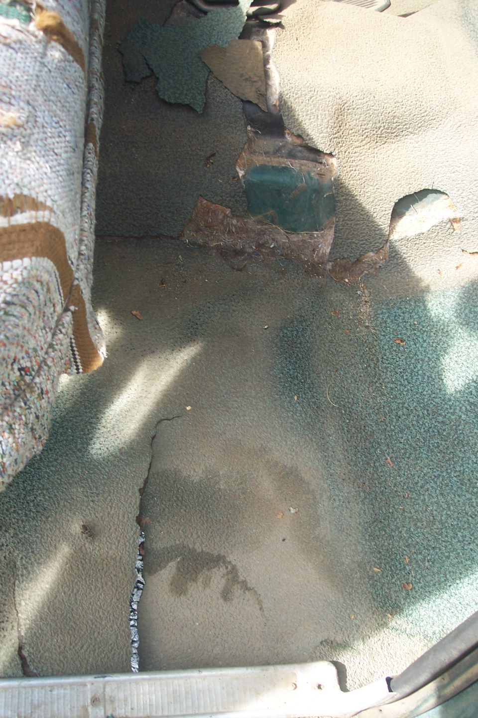Worn interior floor carpet with torn fabric revealing metal and green underlayer. Seat fabric visible on left