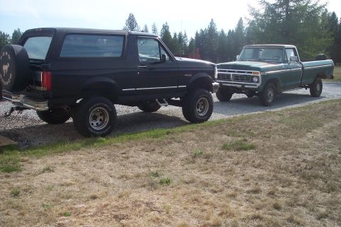 1977 Ford F-150 - Two Ford trucks, a black Bronco and a green F-150, are parked outdoors on a gravel driveway.