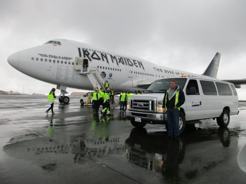 1996 Ford Bronco - A white Ford van is parked on a wet airport tarmac next to a Boeing 747 airplane with 'Iron Maiden' written on its side.
