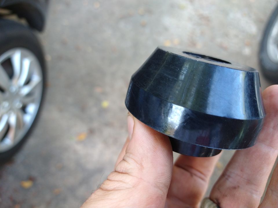 Hand holding a black rubber suspension bushing, with a car wheel in the background