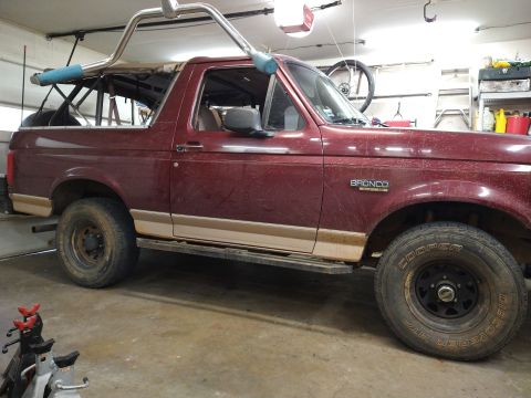 1996 Ford Bronco - The side profile of a maroon Ford Bronco is shown in a garage. The vehicle has a tan lower body trim and a roll cage visible over the inter…