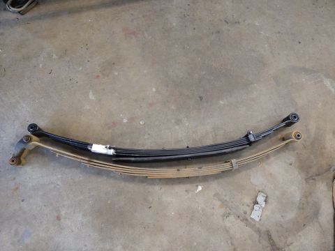 1996 Ford Bronco - Two leaf springs are laid out on a concrete floor. The top leaf spring is black and appears to be newer, while the bottom leaf spring is ru…