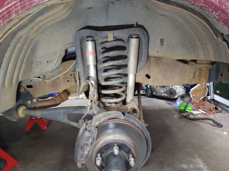 Rear suspension of a red vehicle with coil spring, shock, brake rotor, and caliper, supported by jack stands.
