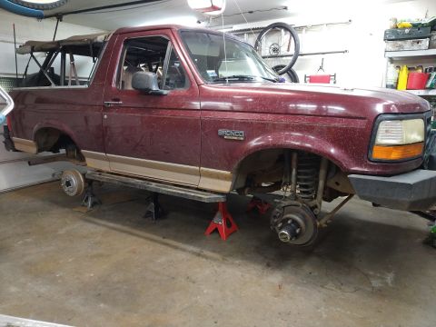 1996 Ford Bronco - Maroon two-tone Ford Bronco raised on jack stands in a garage, front wheels removed exposing the suspension and brakes.