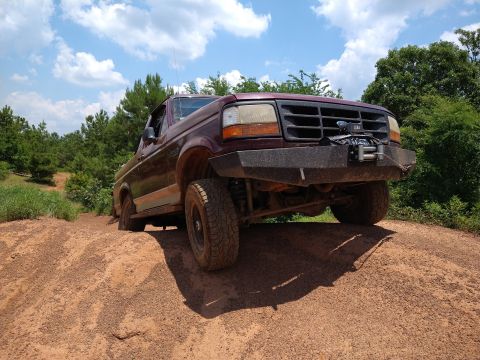 1996 Ford Bronco - A maroon Ford Bronco is shown from a low angle, driving up a dirt hill with trees and a blue sky with clouds in the background. The front o…