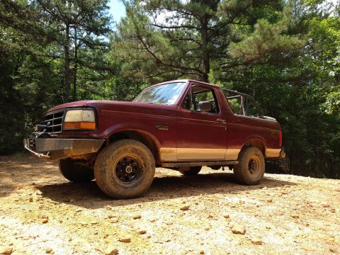 1996 Ford Bronco - A maroon 1996 Ford Bronco with a custom aftermarket bumper sits on a dirt road surrounded by trees. The vehicle is a two-door model with th…