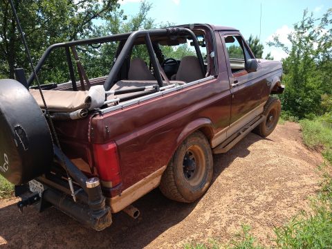 1996 Ford Bronco - A maroon 1990s Ford Bronco is parked on a dirt hill with trees in the background. The vehicle has a roll cage, a spare tire mounted on the…