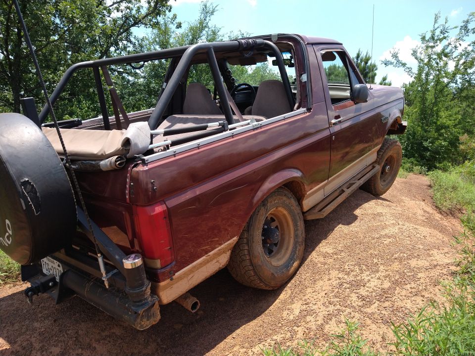 Maroon 1990s Ford Bronco with roll cage and spare tire on a dirt hill.