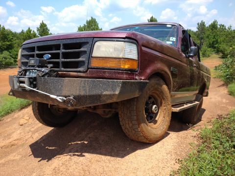1996 Ford Bronco - A maroon Ford Bronco is parked on a dirt road with trees in the background. The front of the vehicle is equipped with a winch mounted on a…