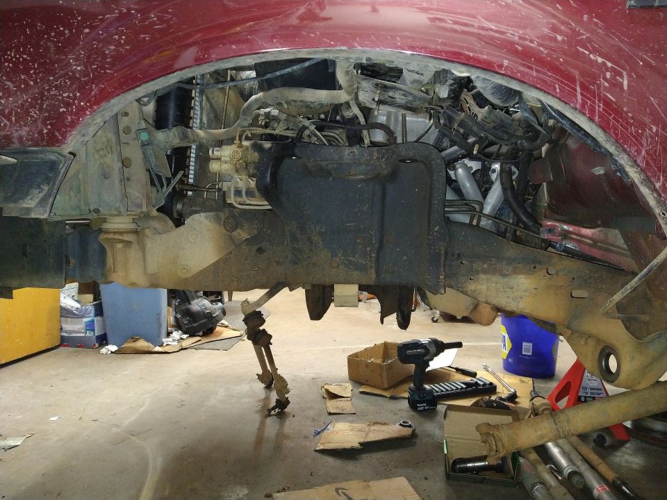 Underside of a red Ford Bronco with suspension and engine parts exposed, tools on the floor.