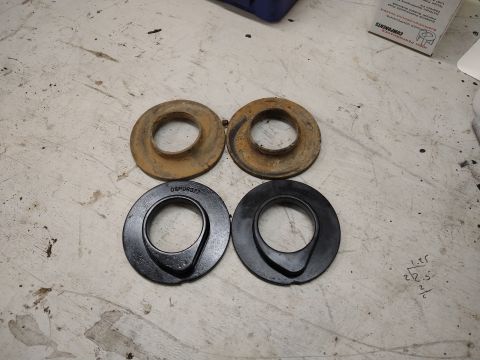 1996 Ford Bronco - Two pairs of suspension spring spacers are laid out on a textured surface. The top pair are rusty metal, while the bottom pair are black.