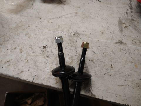 1996 Ford Bronco - Two black suspension components, each with a threaded bolt and a nut, are shown resting on a light-colored, textured surface. One nut is si…