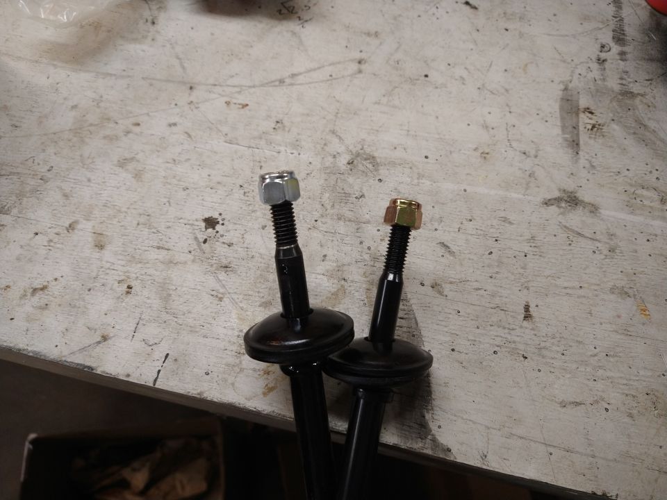 Two black suspension parts with bolts and nuts on a textured surface.