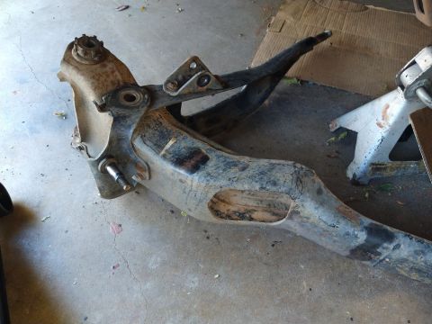 1996 Ford Bronco - A heavily rusted and weathered suspension control arm assembly is laid out on a concrete floor, with a car jack visible in the background.