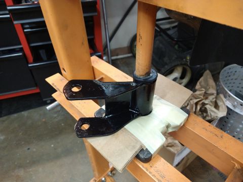 1996 Ford Bronco - A black metal suspension part with two holes is being pressed into place by an orange hydraulic press. The part is resting on cardboard and…