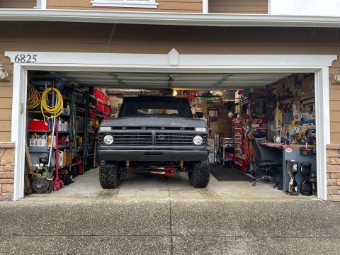 1978 Ford Bronco - A dark gray, lifted 1970s Ford pickup truck is parked inside an open garage. The garage is filled with shelves of tools, supplies, and othe…