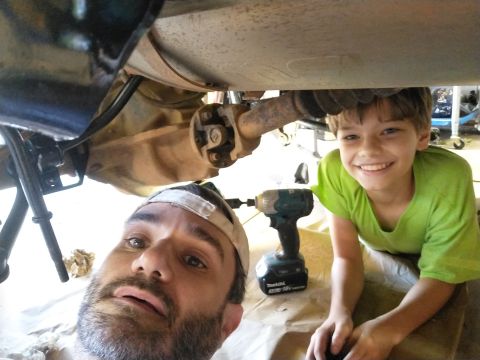1996 Ford Bronco - A father and son are working on the underside of a vehicle, possibly a 1996 Ford Bronco, with a power drill visible.