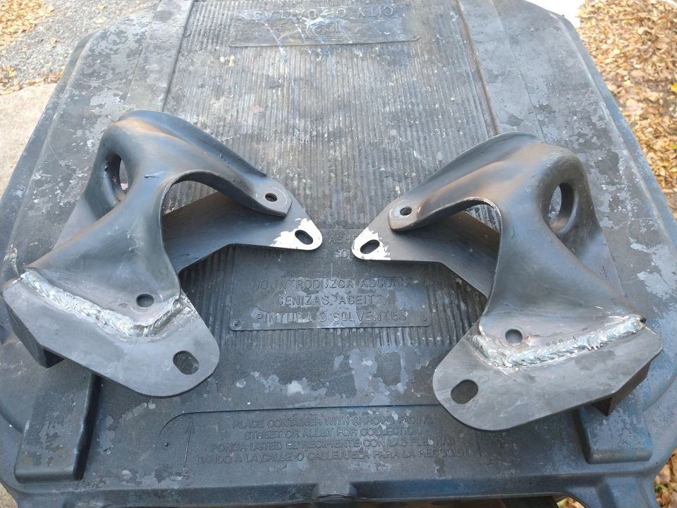 Two black metal automotive brackets with welds on a dark gray surface.
