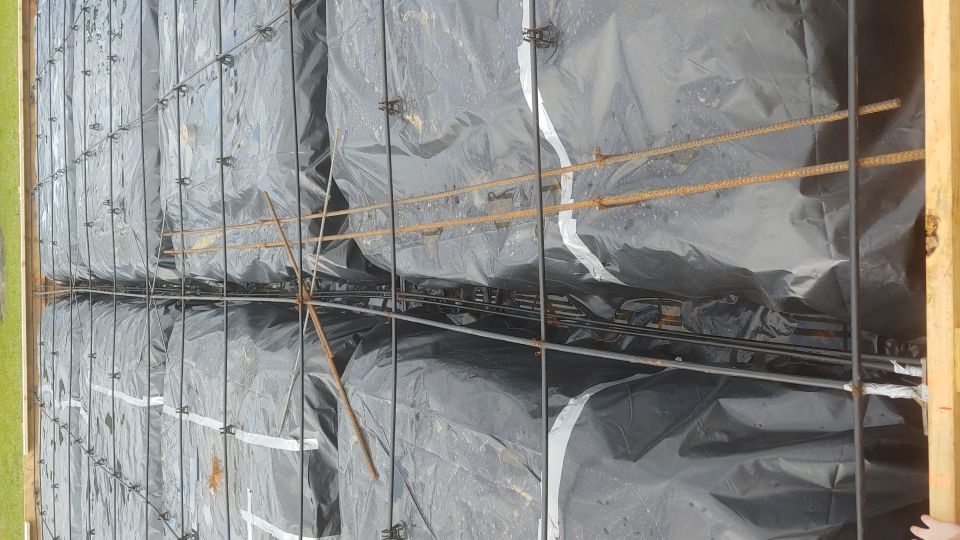 Construction site showing rebar grid covered with black plastic sheeting inside wooden forms