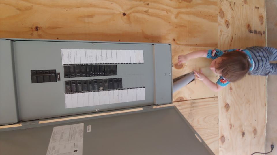 Electrical panel with circuit breakers mounted on a plywood wall, a child is interacting with a pipe entering the panel