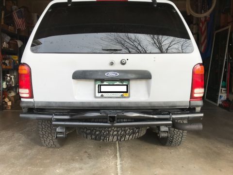 2000 Ford Explorer - The rear view of a white Ford Explorer SUV is shown, featuring a black aftermarket bumper with a trailer hitch and dual exhaust pipes. A sp…