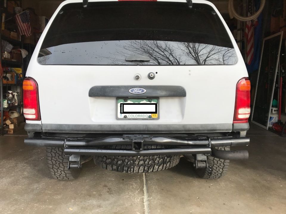 White Ford Explorer rear view with aftermarket bumper, trailer hitch, dual exhaust, and spare tire.